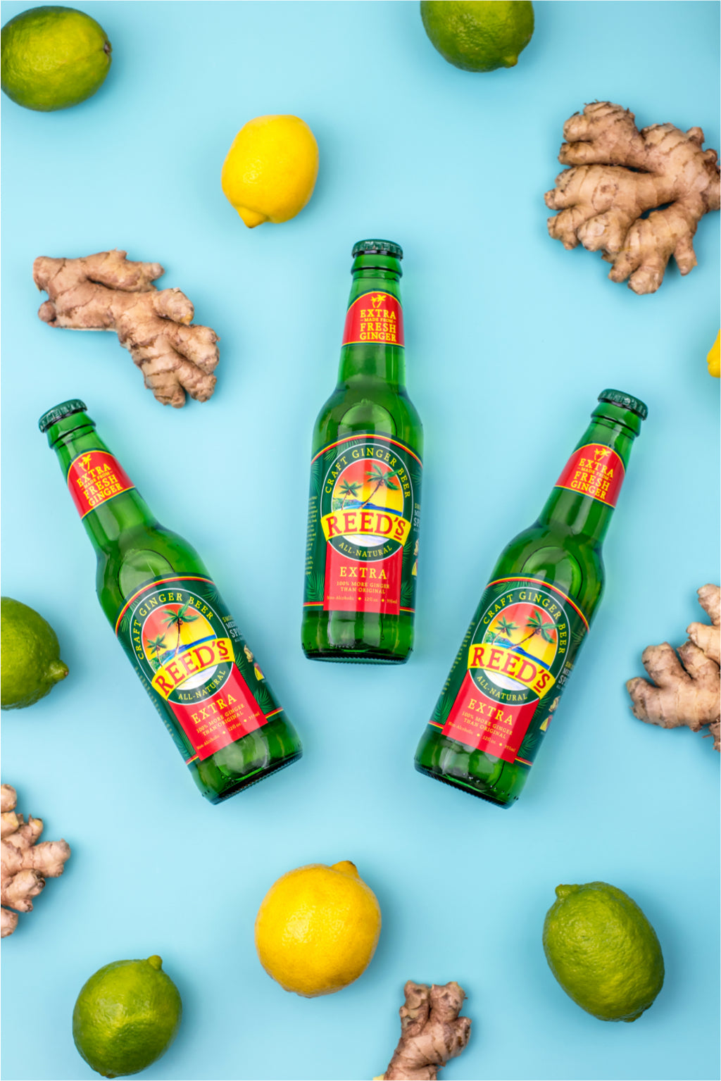 Three glass bottles of Reed's Extra Ginger Beer on a blue background next to pieces of ginger, lemons, and limes.