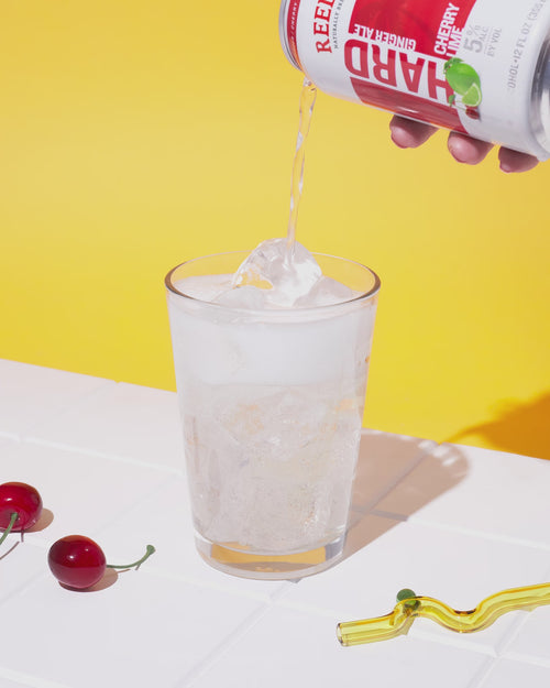 Someone pouring a can of Reed's Hard Ginger Ale into a glass next to cherries and a yellow straw.